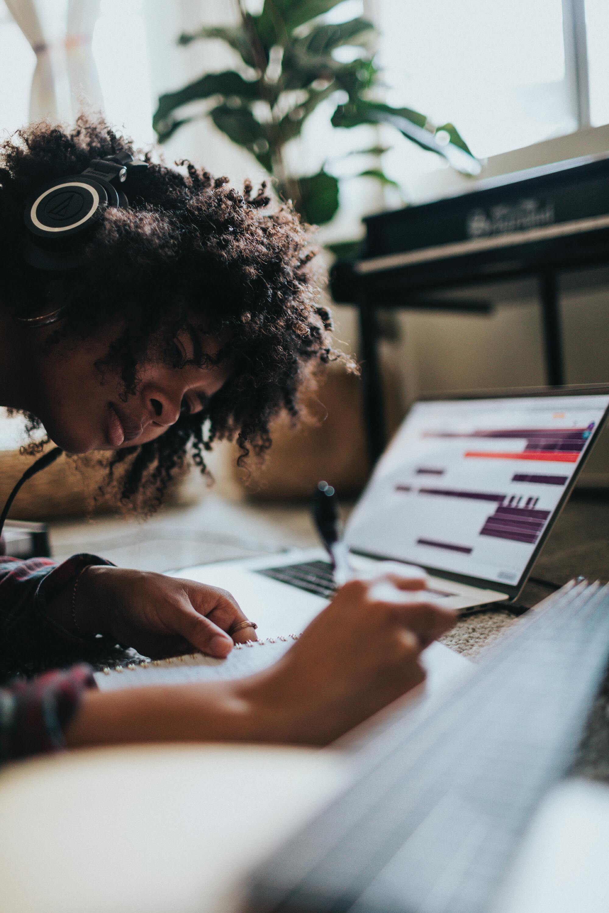Photo by Soundtrap, from Un-Splash. A Black woman with dark afro hair wearing headphones writing intently in a notebook.  There is an open laptop in the background slightly out of focus.