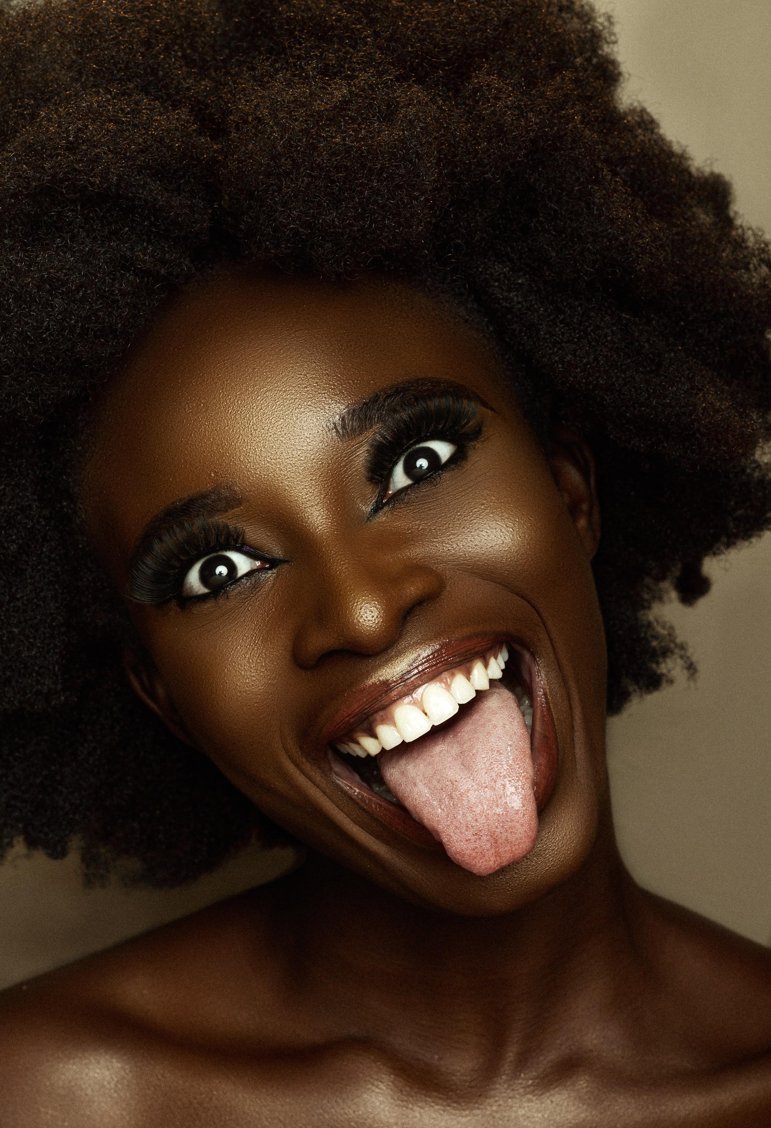 A dark brown Black woman with a large afro is smiling at the camera with her tongue out in a silly way.