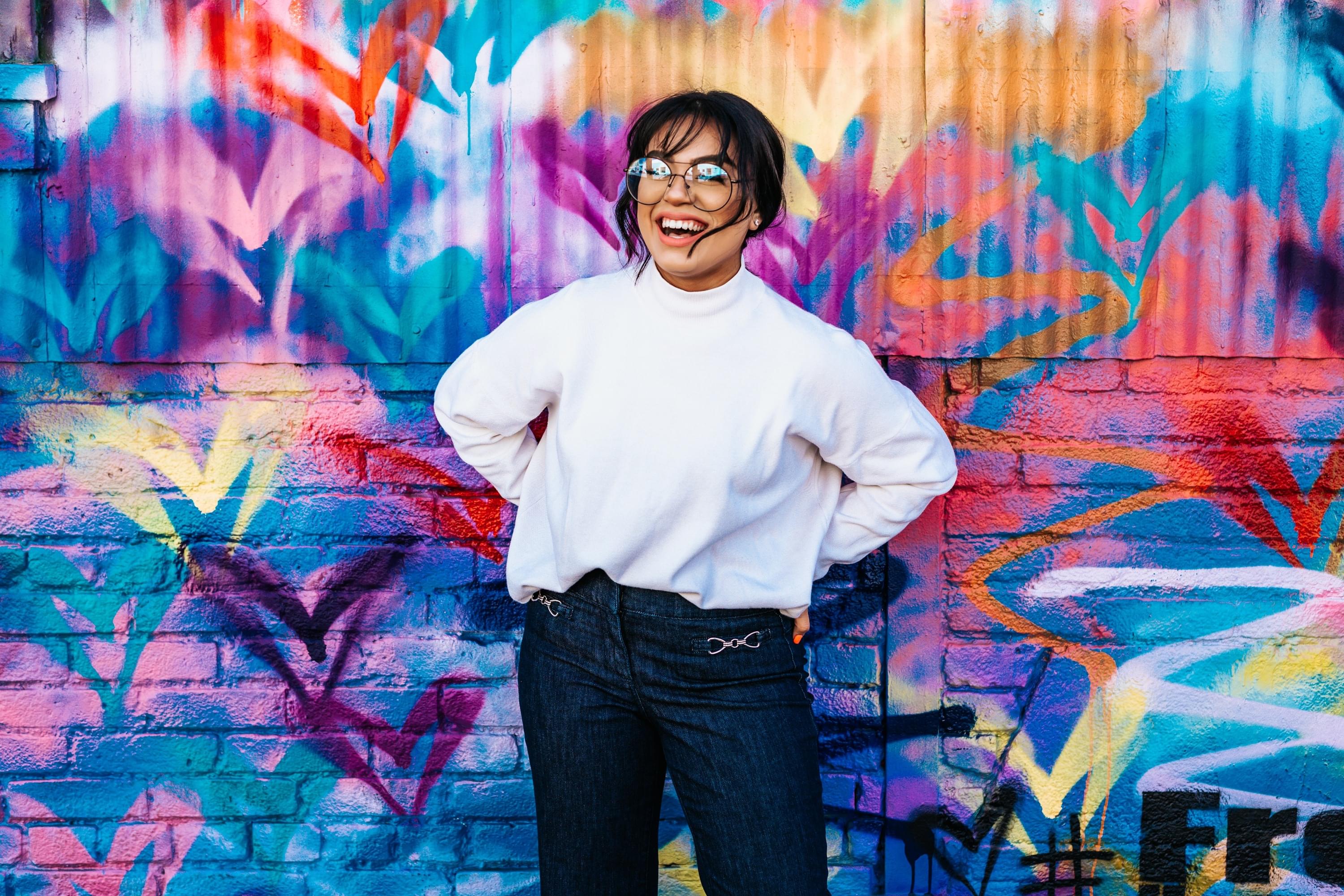 A light brown person with glasses in a white long sleeved top and dark denim jeans smiling and standing against a multicolored mural painted background wall.