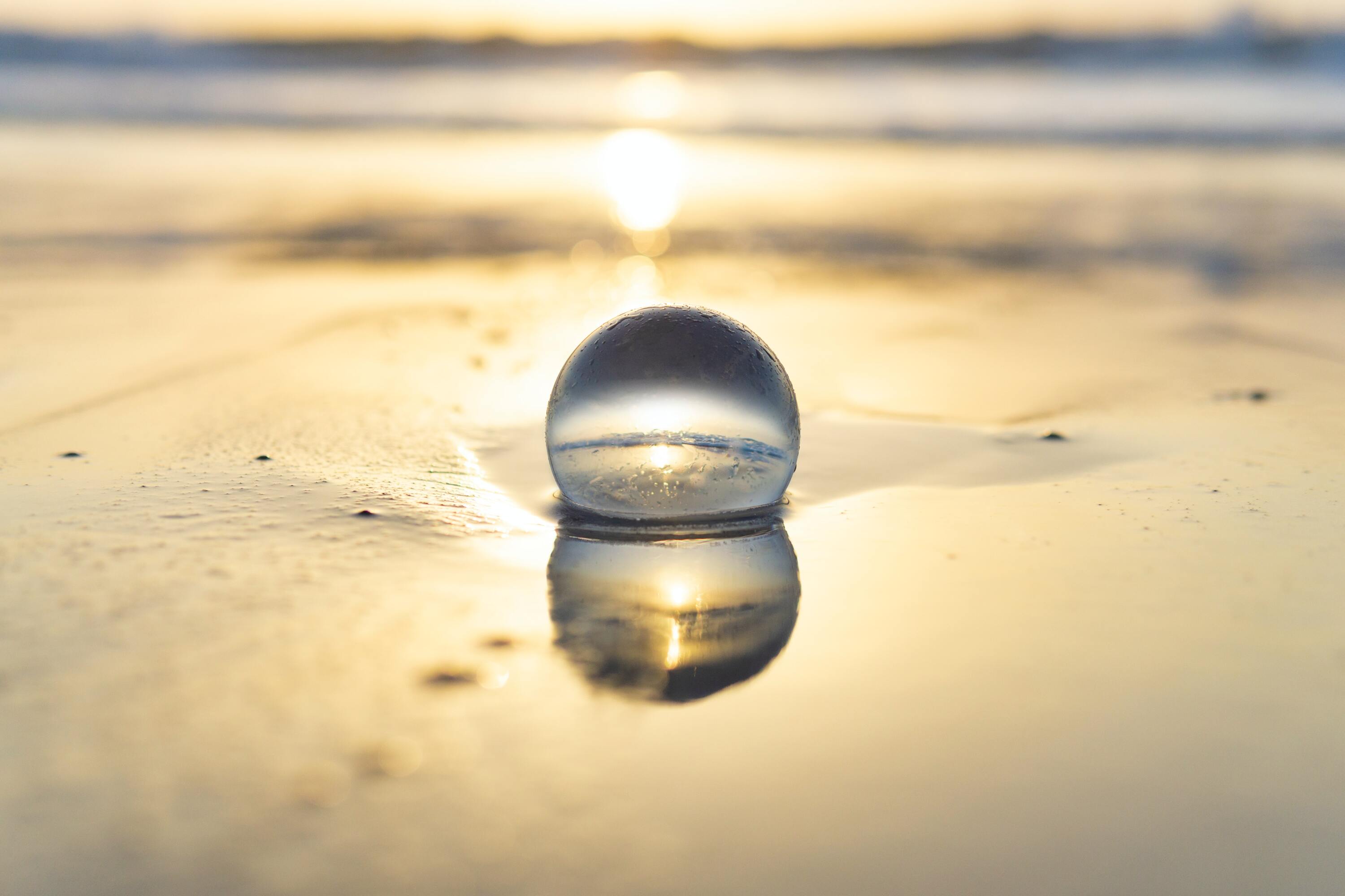 Photo by Joshua Woroniecki, from Un-splash.  On a soft golen background, reflecting the sun, a large clear marble sits in wet sand, reflecting the water within it.